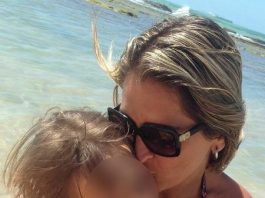 Mãe abraçando e beijando o rosto de uma criança. Eles estão numa praia e o rosto da criança está desfocado.